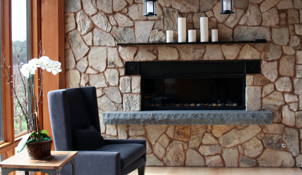 closeup of a beautiful fireplace insert surrounded by cut stone veneer; the fireplace insert has a black wrought-iron mantel and a concrete footer for additional seating close to the fire; decorative candles sit atop the mantel; industrial lights hang from red wood posts above the fireplace