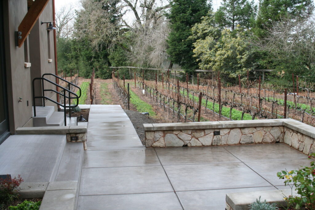 a concrete patio leads around the outside of the winery; the patio is enclosed by low walls featuring cut stone veneer; the vineyard is viewable from the patio