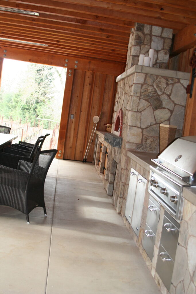 outdoor kitchen features a pizza oven and built-in grill; the counter is made from poured concrete; the cabinets are set into cut stone veneer