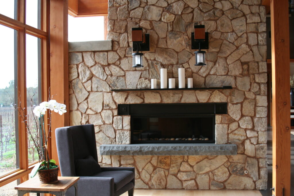 closeup of a beautiful fireplace insert surrounded by cut stone veneer; the fireplace insert has a black wrought-iron mantel and a concrete footer for additional seating close to the fire; decorative candles sit atop the mantel; industrial lights hang from red wood posts above the fireplace