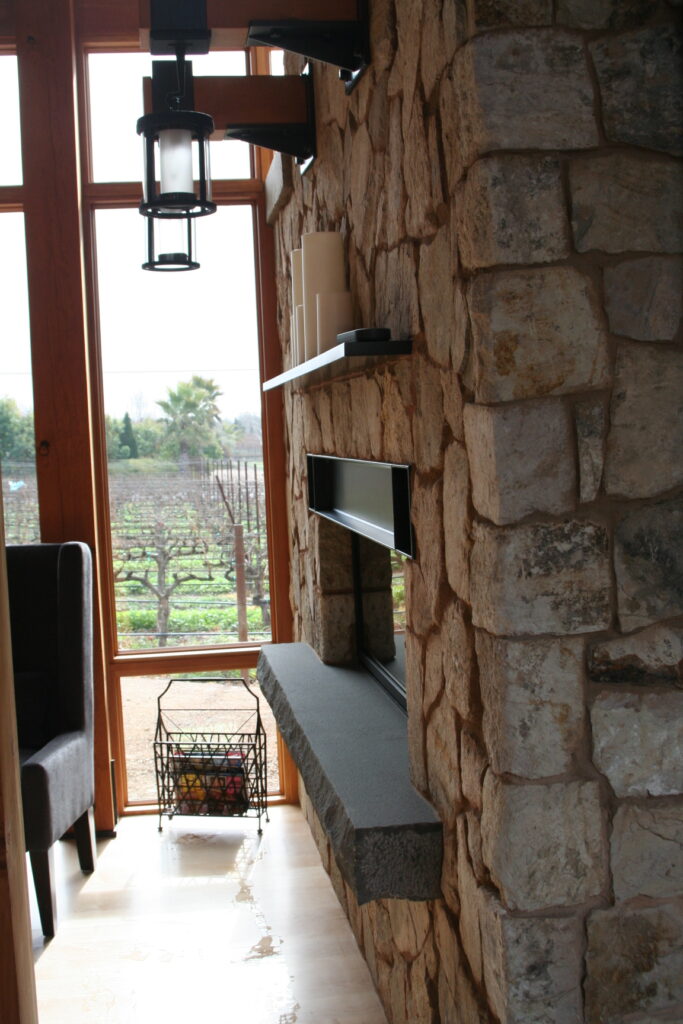 closeup of a beautiful fireplace insert surrounded by cut stone veneer; the fireplace insert has a black wrought-iron mantel and a concrete footer for additional seating close to the fire; decorative candles sit atop the mantel; industrial lights hang from red wood posts above the fireplace