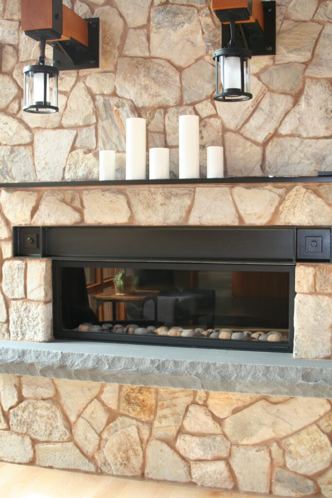 closeup of a beautiful fireplace insert surrounded by cut stone veneer; the fireplace insert has a black wrought-iron mantel and a concrete footer for additional seating close to the fire; decorative candles sit atop the mantel; industrial lights hang from red wood posts above the fireplace