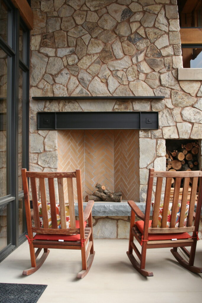 closeup of the interior of an outdoor fireplace; the interior of the fireplace features bricks laid in a herringbone pattern; two rocking chairs rest in front of the fireplace with festive pillows