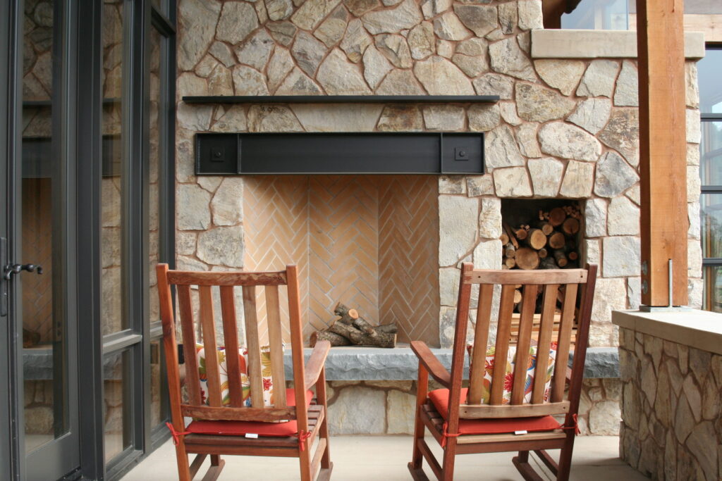 closeup of the interior of an outdoor fireplace; the interior of the fireplace features bricks laid in a herringbone pattern; two rocking chairs rest in front of the fireplace with festive pillows