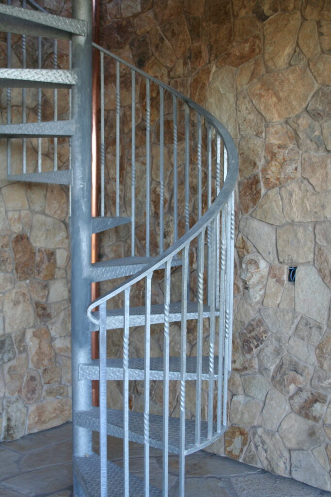 an exterior steel spiral staircase rests against the stone veneer of the residence; you can see a copper utility pipe in the background