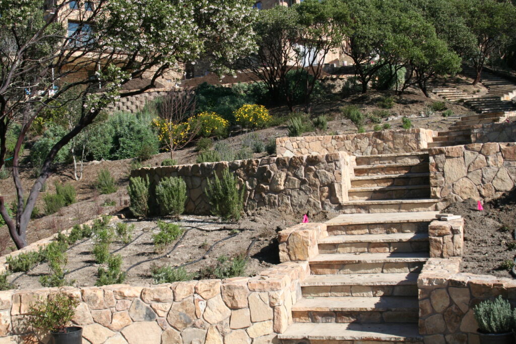 cut stone steps with single stone treads lined on either side by cut stone walls leads up a hill through a tiered garden with exposed irrigation