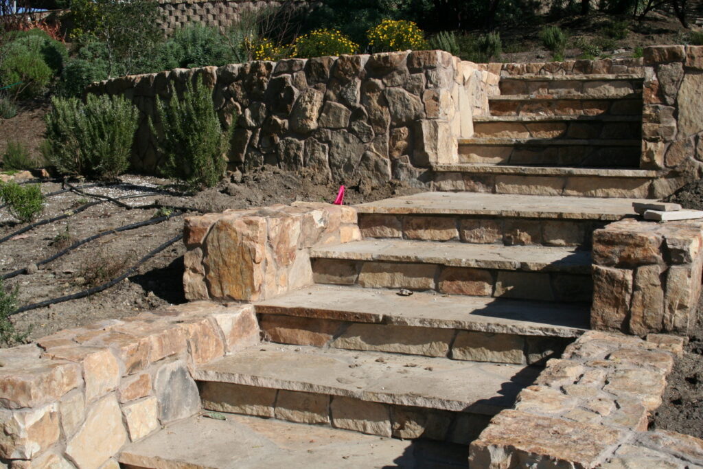 cut stone steps with single stone treads lined on either side by cut stone walls leads up a hill through a tiered garden with exposed irrigation