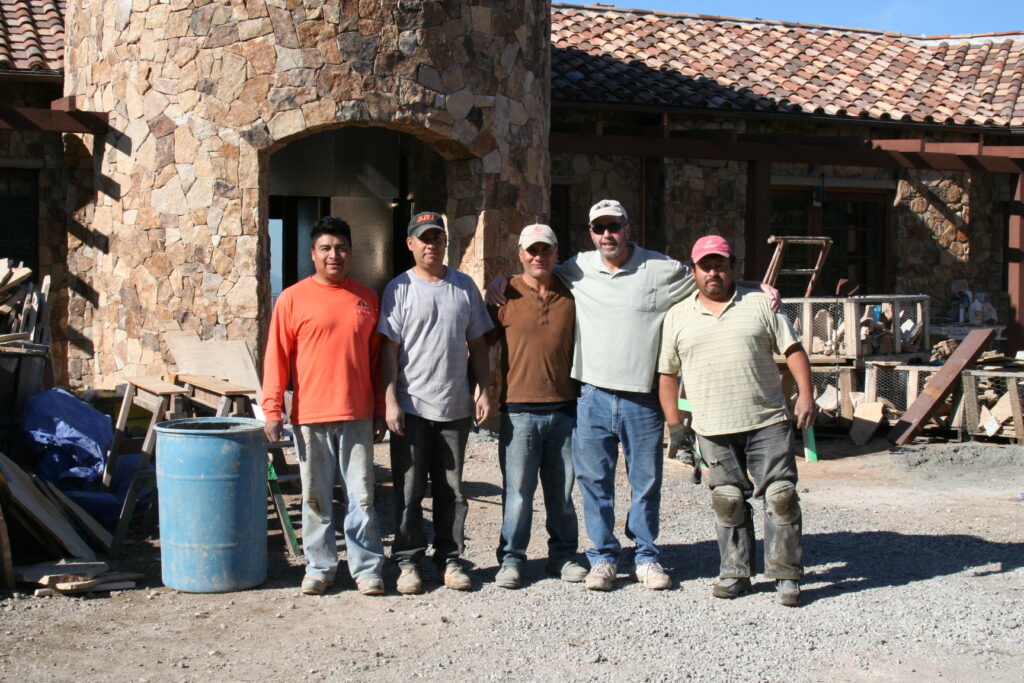 Conrad poses with his professional masonry team outside the front door of an estate still under construction; the facade of the building is covered in cut stone veneer