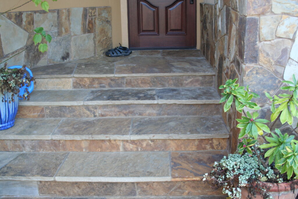 cut stone skirting blends into the front landing with steps of a Santa Rosa residence; a pair of athletic sandals sit outside the dark wooden front door