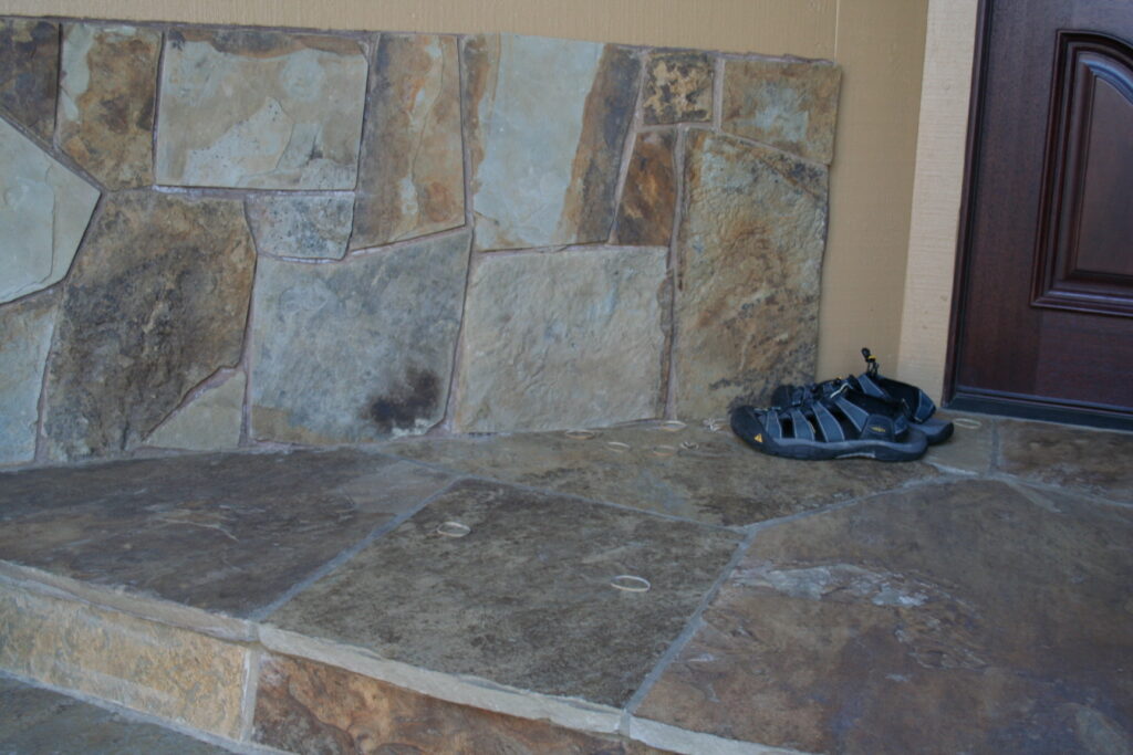 cut stone skirting blends into the front landing with steps of a Santa Rosa residence; a pair of athletic sandals sit outside the dark wooden front door