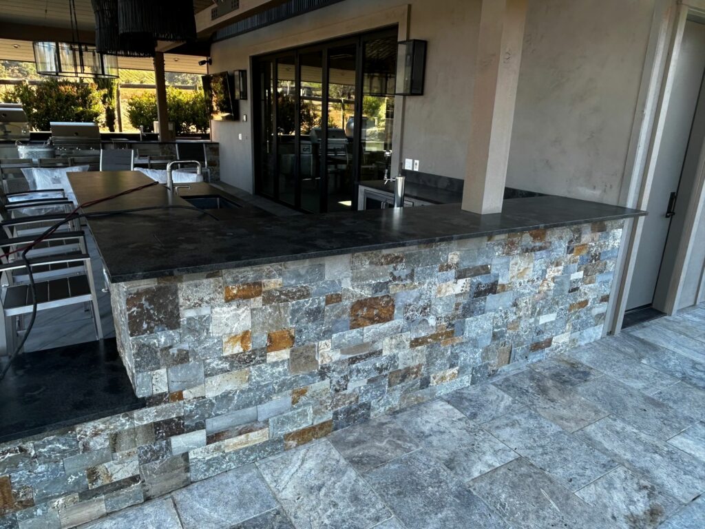 outdoor kitchen with 2 built-in grills; the counters are supported by stone walls; the patio just outside the outdoor kitchen is laid with large stone tiles set in a brickwork pattern