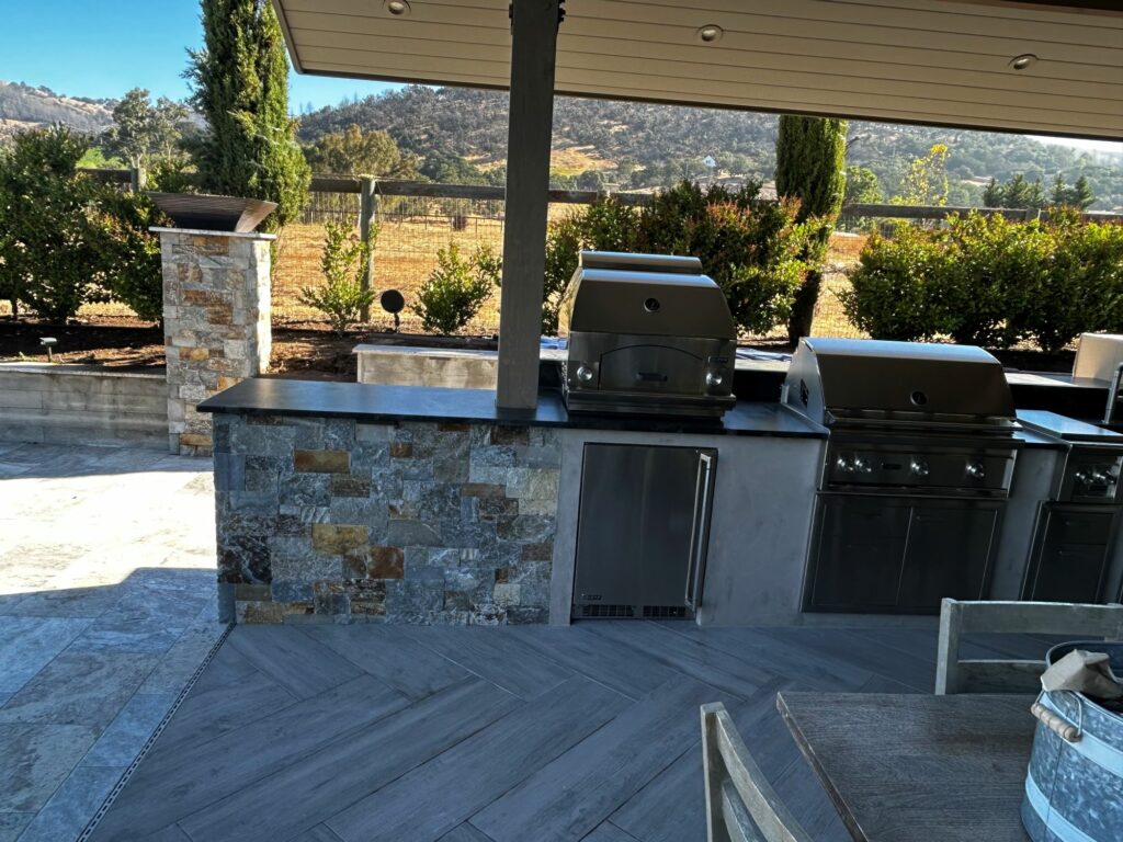 outdoor kitchen with 2 built-in grills; the counters are supported by stone walls; the floor of the outdoor kitchen is laid with stone tiles set in a herringbone pattern