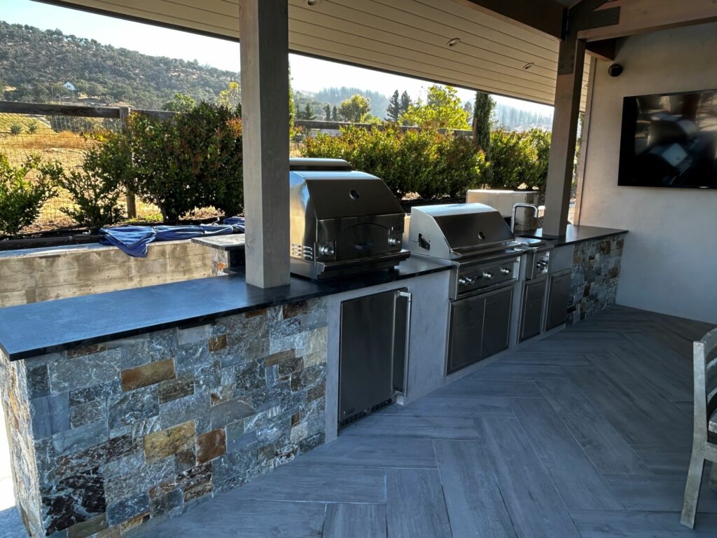 outdoor kitchen with 2 built-in grills; the counters are supported by stone walls; the floor of the outdoor kitchen is laid with stone tiles set in a herringbone pattern