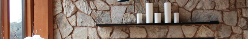 closeup of a beautiful fireplace insert surrounded by cut stone veneer; the fireplace insert has a black wrought-iron mantel and a concrete footer for additional seating close to the fire; decorative candles sit atop the mantel; industrial lights hang from red wood posts above the fireplace